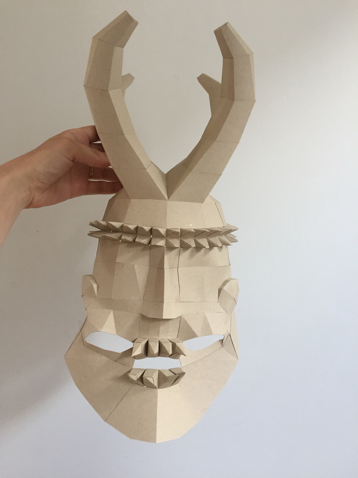 Unpainted full geometric horned mask prototype held against a wall